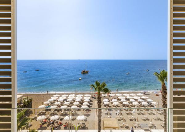 Spiaggia soleggiata con ombrelloni bianchi, mare azzurro e barche lontane.
